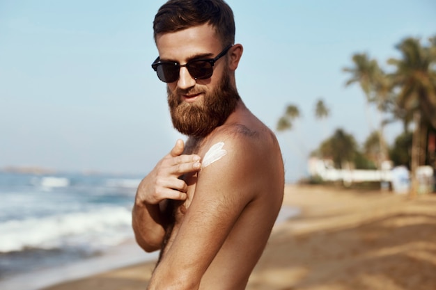 Uomo bello con la barba, in occhiali da sole, prendere il sole con crema solare corpo in estate.