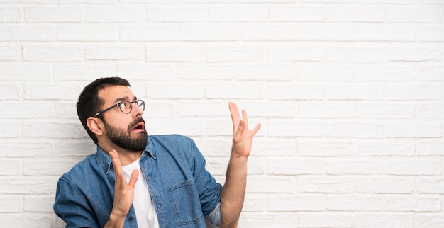 Uomo bello con la barba sopra il muro di mattoni bianco nervoso e spaventato