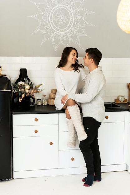 Uomo bello con la moglie incinta che si abbraccia in cucina. la ragazza si siede sul tavolo e abbraccia il marito in piedi con un sorriso stampato in faccia.