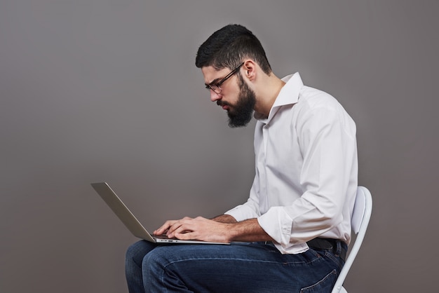 Uomo bello di affari in vetri e vestito che tiene il computer portatile in mano e scrivere qualcosa. Vista laterale. Grigio isolato