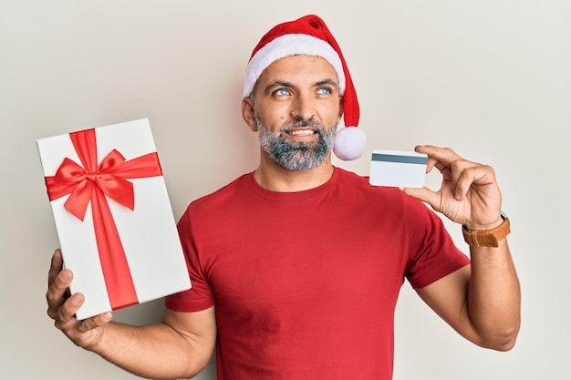 Uomo bello di mezza età che indossa il cappello di crismast che tiene il regalo e la carta di credito sorridendo guardando di lato e fissando lontano pensando.