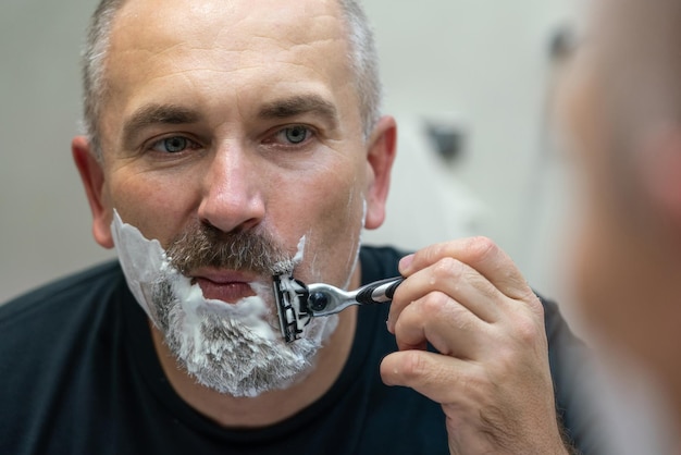 Uomo bello di mezza età che si rade la barba in bagno