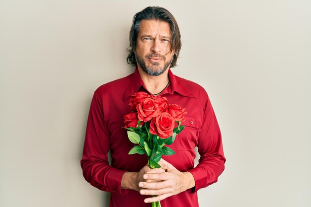 Uomo bello di mezza età che tiene il mazzo di rose rosse concetto di dubbio espressione confusa e all'oscuro