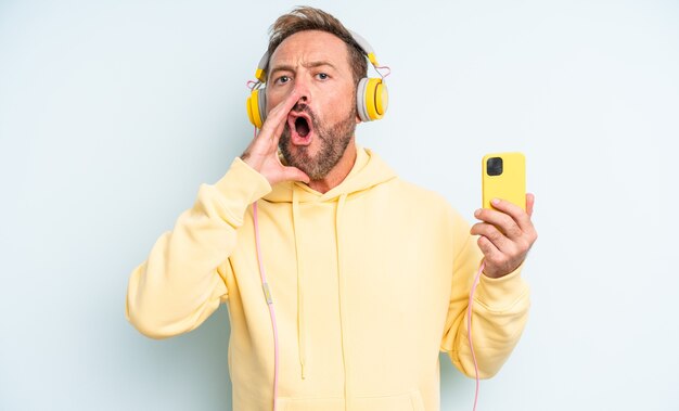 Uomo bello di mezza età sentirsi felice, dando un grande grido con le mani vicino alla bocca. cuffie e concetto di smartphone