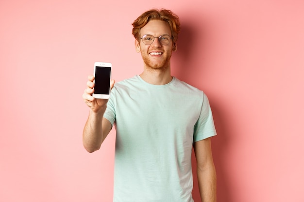 Uomo bello di redhead in vetri che mostrano lo schermo in bianco dello smartphone e sorridente, dimostrano la promozione o l'applicazione in linea, stando sopra il fondo rosa