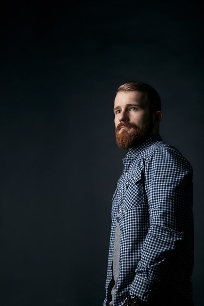 Uomo bello ed elegante con la barba rossa pensierosa in camicia a scacchi blu, ritratto in studio su sfondo scuro