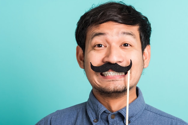 Uomo bello felice asiatico del ritratto che propone che tiene una carta divertente dei baffi o baffi falsi dell'annata sulla sua bocca