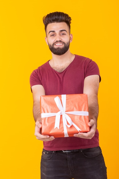 Uomo bello giovane hipster carino in camicia bordeaux che tiene una confezione regalo rossa in posa su un giallo