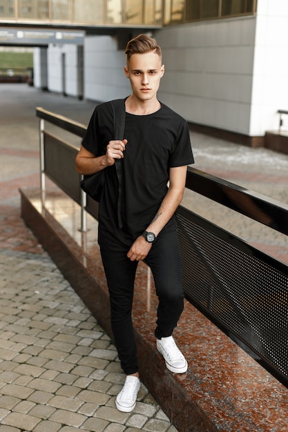 Uomo bello hipster in maglietta nera alla moda, pantaloni e scarpe da ginnastica bianche con uno zaino in piedi vicino all'edificio moderno