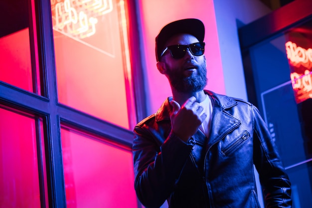 Uomo bello hipster per le strade della città illuminata da insegne al neon. Indossa una giacca da motociclista in pelle o una giacca con zip asimmetrica con berretto nero, jeans e occhiali da sole.