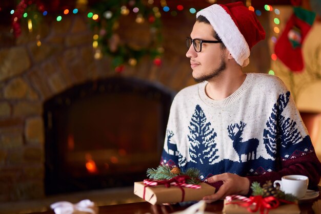 Uomo bello in cappello della Santa che beve la creazione di regali Bellissimo sfondo con caminetto Posto accogliente