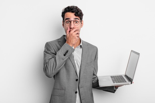 Uomo bello ispanico che copre la bocca con le mani con uno scioccato. concetto di business e laptop