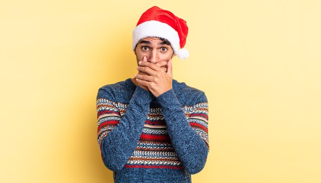 Uomo bello ispanico che copre la bocca con le mani con uno scioccato. concetto di natale