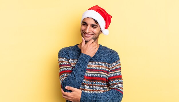 Uomo bello ispanico che sorride con un'espressione felice e sicura con la mano sul mento. concetto di natale