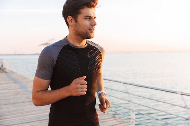 Uomo bello muscolare in tuta da jogging lungo il molo o il lungomare in riva al mare, durante l'alba