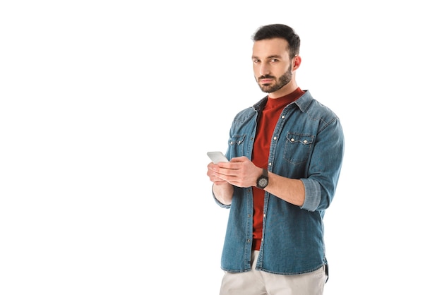 Uomo bello pensieroso in camicia di jeans utilizzando smartphone isolato su bianco