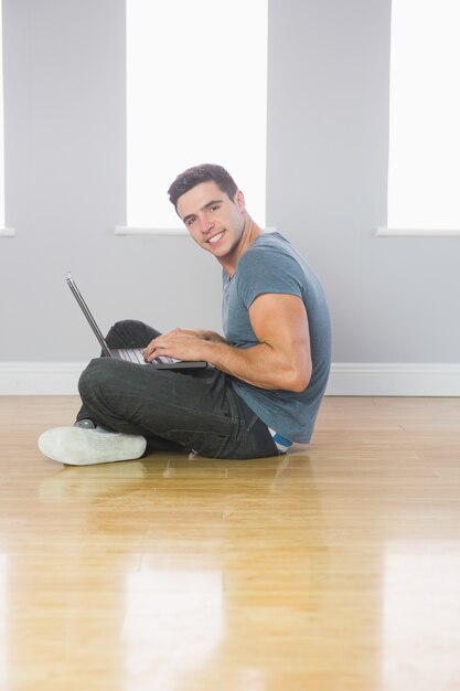 Uomo bello sorridente che per mezzo del computer portatile che si siede sul pavimento