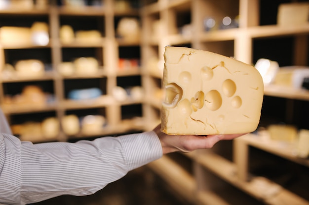 Uomo bello tenere in mano grande fetta di formaggio maasdam. Formaggio con grandi buchi. Sfondo di scaffali con formaggio