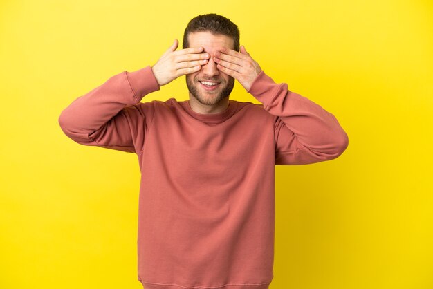 Uomo biondo bello sopra gli occhi gialli isolati della copertura murale con le mani e sorridente