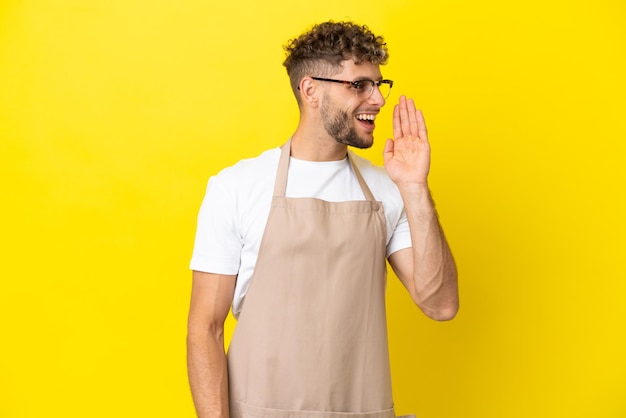 Uomo biondo cameriere ristorante isolato su sfondo giallo che grida con la bocca spalancata lateralmente