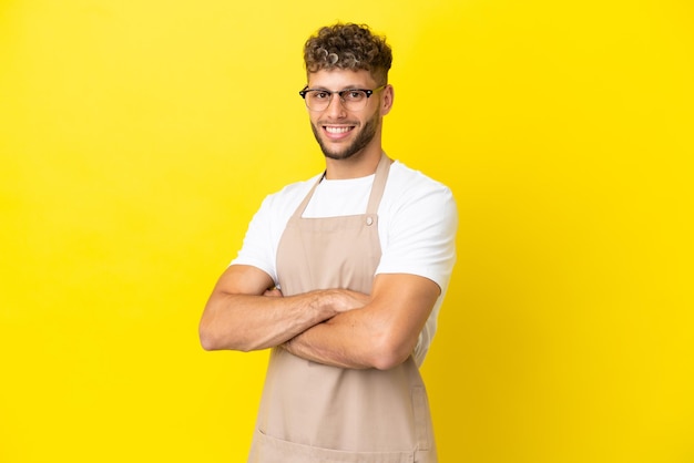 Uomo biondo cameriere ristorante isolato su sfondo giallo con le braccia incrociate e guardando avanti