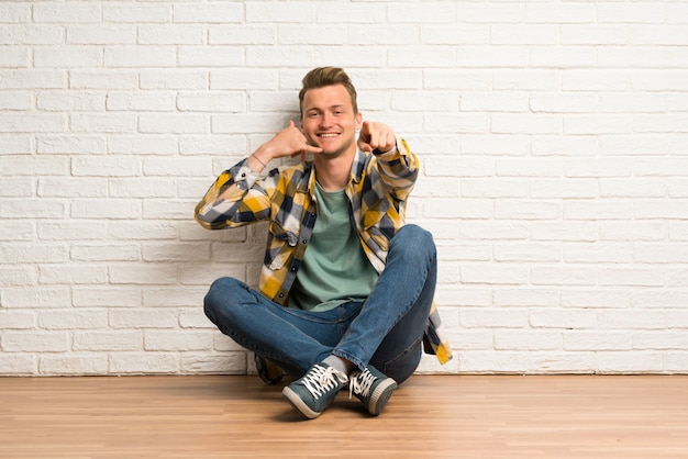 Uomo biondo che si siede sul pavimento che fa gesto del telefono e che indica fronte