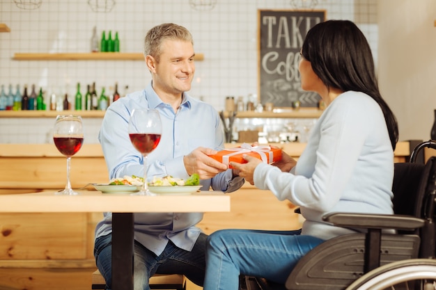 Uomo biondo contenuto bello che sorride e che dà un regalo alla sua donna disabile piuttosto gioiosa mentre cena