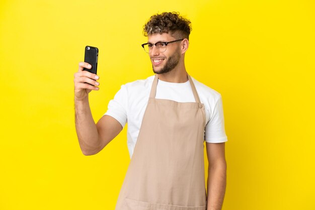 Uomo biondo del cameriere del ristorante isolato su fondo giallo che fa un selfie