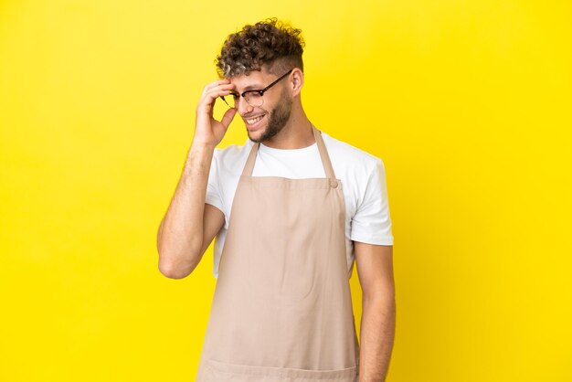 Uomo biondo del cameriere del ristorante isolato su fondo giallo che ride