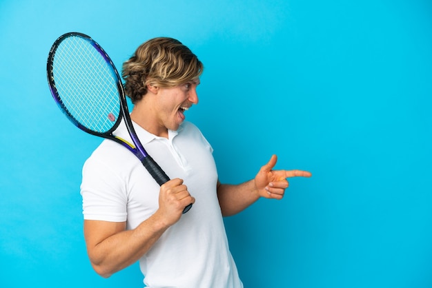 Uomo biondo del giocatore di tennis isolato sulla parete blu che indica il dito di lato e che presenta un prodotto