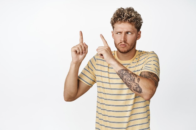 Uomo biondo sospettoso con i capelli ricci che puntano e guardano l'angolo in alto a sinistra con la faccia incredula dubbiosa in piedi in abiti casual su sfondo bianco