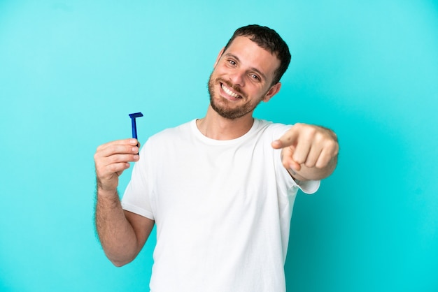 Uomo brasiliano che si rade la barba isolato su sfondo blu che punta davanti con espressione felice