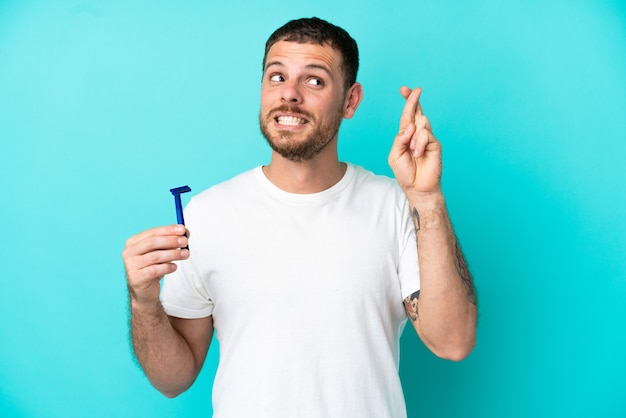Uomo brasiliano che si rade la barba isolato su sfondo blu con le dita incrociate e augurando il meglio