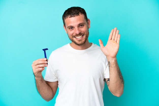 Uomo brasiliano che si rade la barba isolato su sfondo blu salutando con la mano con espressione felice