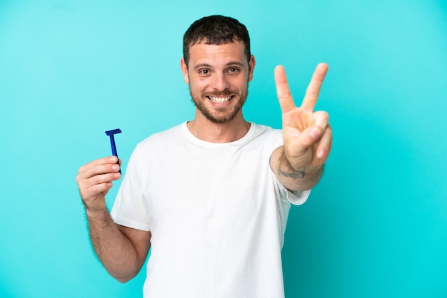 Uomo brasiliano che si rade la barba isolato su sfondo blu sorridendo e mostrando il segno della vittoria