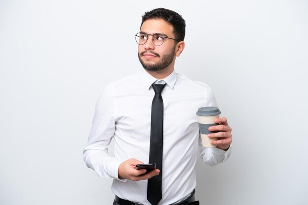 Uomo brasiliano d'affari isolato su sfondo bianco che tiene il caffè da portare via e un cellulare mentre pensa a qualcosa