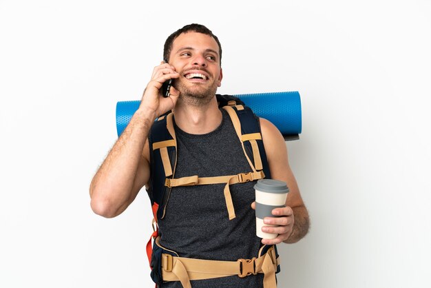 Uomo brasiliano dell'alpinista con un grande zaino sopra fondo bianco isolato che tiene il caffè da portare via e un cellulare
