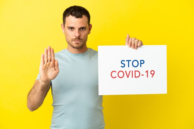 Uomo brasiliano su sfondo viola isolato che tiene un cartello con il testo Stop Covid 19 e fa il segnale di stop