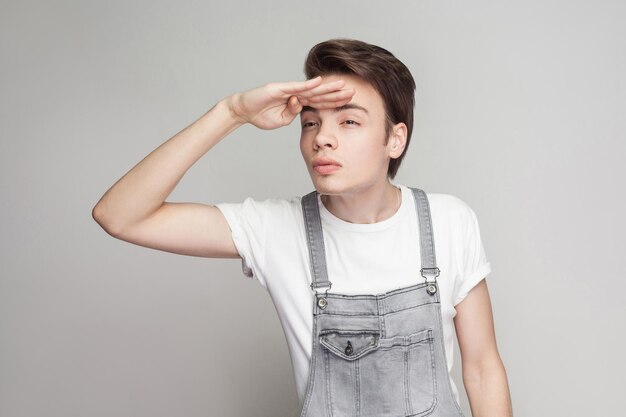 Uomo bruna serio in stile casual con t-shirt bianca e tuta di jeans in piedi tenendosi per mano sulla fronte e guardando lontano con la faccia seria. girato in studio al coperto, isolato su sfondo grigio.