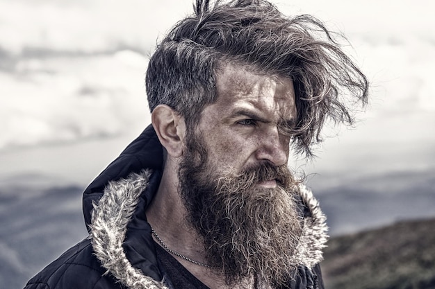 Uomo brutale hipster uomo barbuto barba lunga brutale caucasico con baffi ragazzo con la barba lunga con capelli alla moda ottenere barbe taglio di capelli sulla cima della montagna ventosa sul cielo nuvoloso naturale uomo barbuto maturo