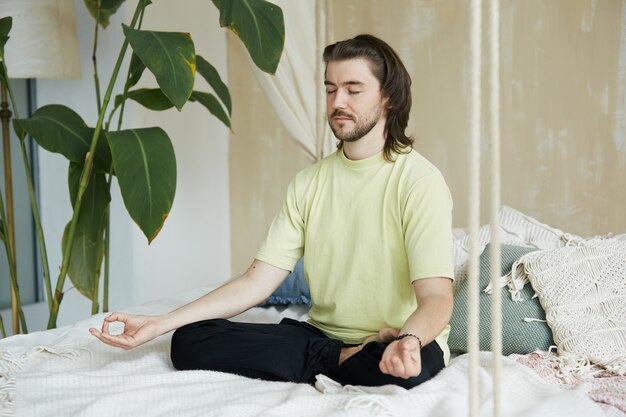 Uomo calmo seduto sul letto con gli occhi chiusi, maschio in lotus asana e yoga mudra che pratica la meditazione sul letto