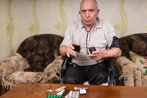 Uomo calvo anziano con bisogni speciali, seduto sulla sedia a rotelle in soggiorno, guardando la telecamera mentre tiene in mano un succo di frutta salutare e un apparecchio per la pressione sanguigna