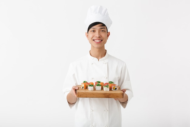 uomo capo cinese positivo in uniforme bianca del cuoco che sorride alla macchina fotografica mentre tiene il piatto con l'insieme dei sushi isolato sopra la parete bianca