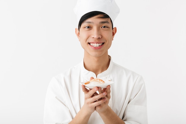 uomo capo cinese soddisfatto in uniforme bianca cuoco sorridendo alla telecamera mentre si tiene piatto con frutti di mare sushi isolato sopra il muro bianco