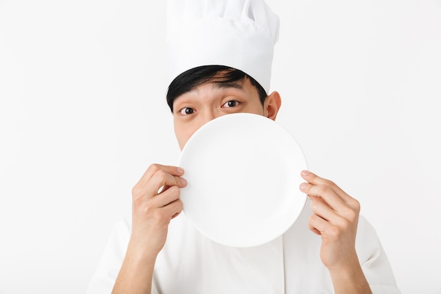 uomo capo divertente asiatico in uniforme bianca del cuoco che sorride alla macchina fotografica mentre tiene un piatto isolato sopra il muro bianco