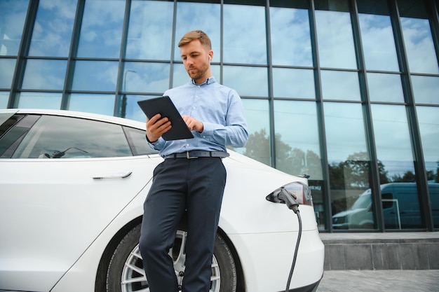 Uomo casual con smartphone vicino all'auto elettrica in attesa della fine del processo di ricarica della batteria