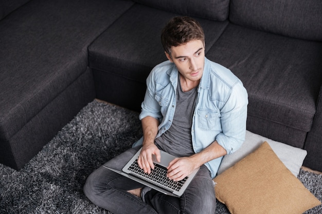 Uomo casual premuroso che lavora al computer portatile mentre è seduto sul tappeto a casa