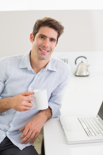 Uomo casuale sorridente con la tazza di caffè con il computer portatile a casa