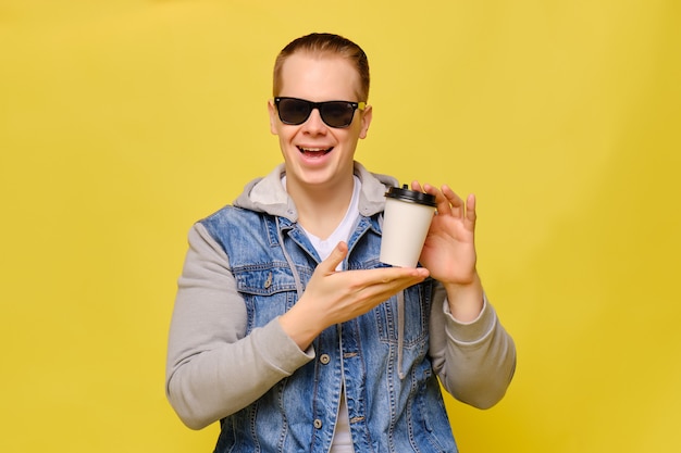 Uomo caucasico alla moda in jeans e occhiali da sole su un fondo giallo. Mani con aria di sfida tiene una tazza di carta usa e getta per il caffè.