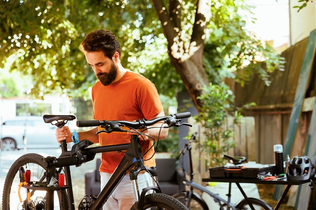 Uomo caucasico atletico che fa la manutenzione annuale della bicicletta mentre usa gli strumenti nel cortile durante l'estate Giovane uomo sportivo che afferra la bicicletta danneggiata per la regolazione sul banco di riparazione